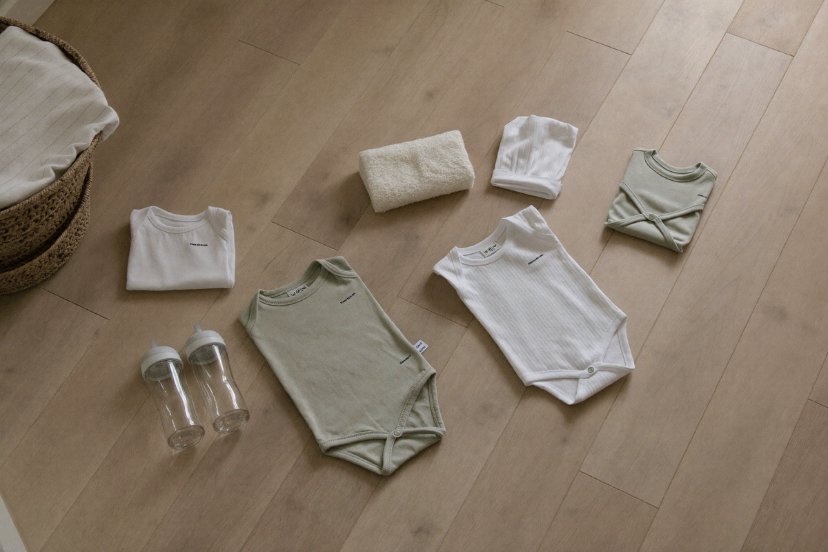 Vue de dessus des essentiels de naissance posés au sol : bodys pliés, biberons, matelas à langer, sortie de bain, gigoteuse Image